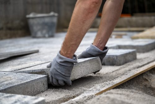 Low angle view of male hands wearing protective gloves placing a cement tile in line with the others while paving the domestic outdoor patio.