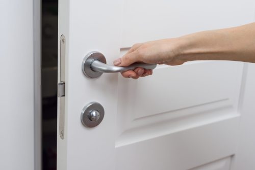 The hand opens the door slightly. Detail of a white interior door with a chrome door handle and latch.